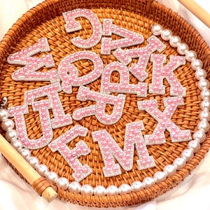 May include: A collection of pink pearl-studded alphabet letters, each outlined with silver rhinestones, arranged in a woven basket. A strand of white pearls encircles the basket's edge, adding a touch of elegance to the decorative letters.