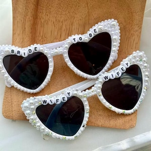 May include: White heart-shaped sunglasses with black lenses. The frames are decorated with pearls and small rhinestones. One pair reads "BRIDE TO BE" and the other reads "WIFEY".