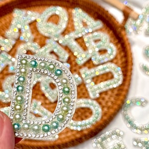 Könnte beinhalten: Nahaufnahme des Buchstabens "D", verziert mit klaren Strasssteinen und hellgrünen Perlen. Der Buchstabe wird vor einem geflochtenen Tablett mit anderen dekorativen Buchstaben gehalten. Die Buchstaben sind hellgrün.