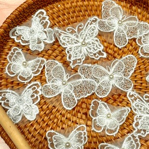 Könnte beinhalten: Weiße Spitzen-Schmetterlingsapplikationen mit Perlen in der Mitte sind auf einem Weidenkorb angeordnet. Die zarten Schmetterlingsflügel weisen komplizierte Muster auf. Einige Schmetterlinge haben einen einzelnen Strassstein. Ideal für Bastelprojekte.