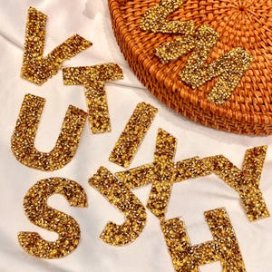 Könnte beinhalten: Goldfarbene Strassbuchstaben mit dem Namen "KATHY" liegen auf einer weißen Oberfläche verstreut. Die Buchstaben sind mit kleinen, funkelnden Strasssteinen verziert. Ein geflochtener Korb ist teilweise im Hintergrund zu sehen.