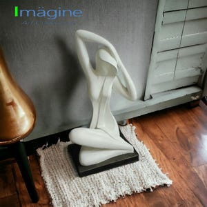 May include: White abstract sculpture of a person sitting with their arms raised above their head. The sculpture is on a black base and is sitting on a white rug.