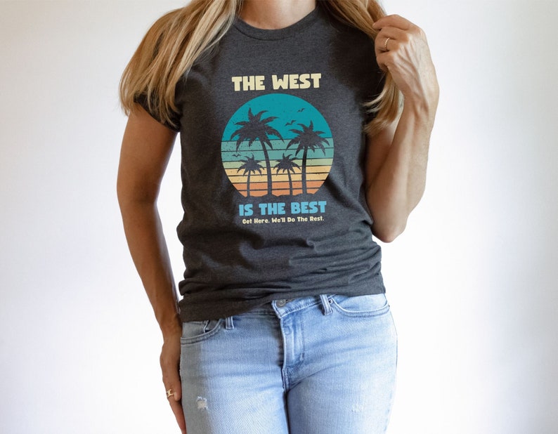 Op de afbeelding: Een donkergrijs T-shirt met een retro zonsondergangafbeelding en palmbomen. De tekst op het shirt luidt "The West is the Best. Get Here, We'll Do The Rest."