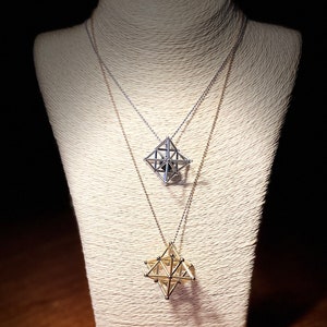 May include: Two geometric pendant necklaces on a white display stand. The top necklace has a silver pendant and the bottom necklace has a gold pendant.