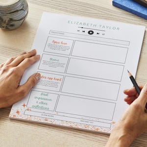 May include: A sheet of white paper with the text "ELIZABETH TAYLOR" and sections for predictions, including "lyric love" and "theme." A pencil is held over the paper, and a mug of coffee sits nearby. The paper has a decorative border.