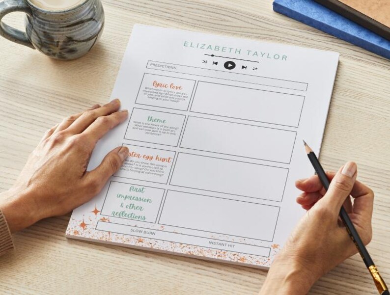 May include: A sheet of white paper with the text "ELIZABETH TAYLOR" and sections for predictions, including "lyric love" and "theme." A pencil is held over the paper, and a mug of coffee sits nearby. The paper has a decorative border.