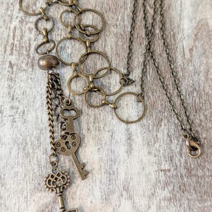 May include: A long gold necklace with a chain of interlocking circles and a dangling charm of multiple keys. The keys are antique-looking and have intricate designs.