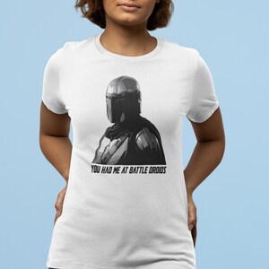 May include: White t-shirt with a black and white image of a Mandalorian character wearing a helmet. The text "You had me at battle droids" is printed below the image.