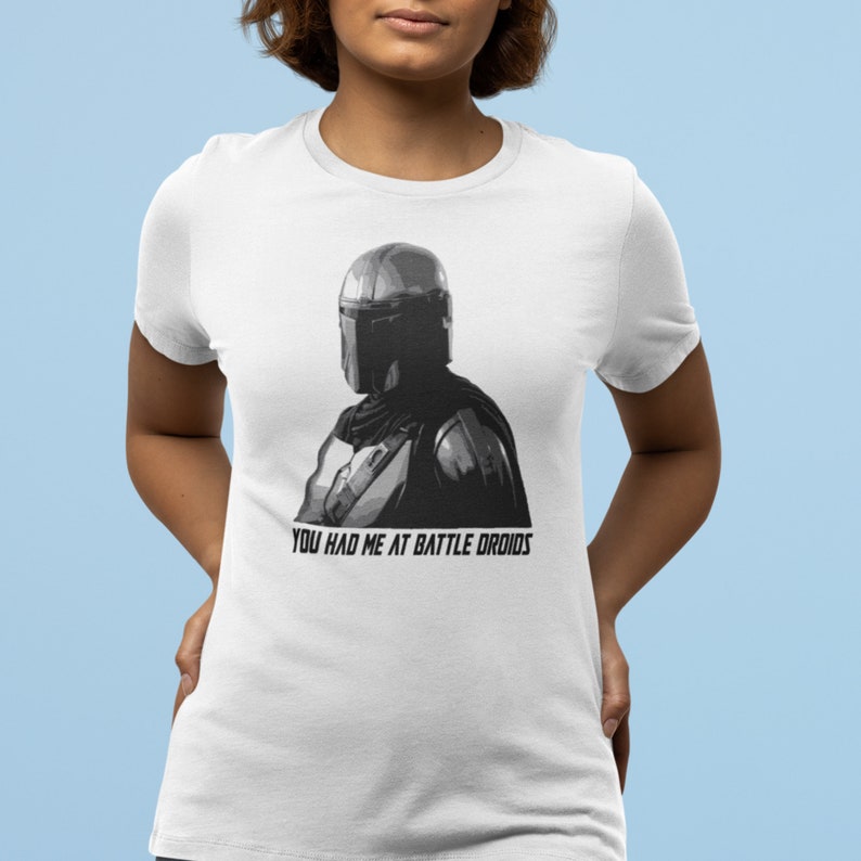 May include: White t-shirt with a black and white image of a Mandalorian character wearing a helmet. The text "You had me at battle droids" is printed below the image.