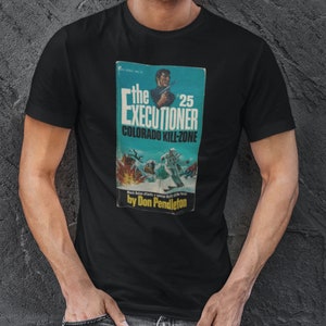 May include: Black t-shirt with a vintage paperback book cover graphic. The book cover features a man holding a gun and the title "The Executioner #25 Colorado Kill-Zone" by Don Pendleton.