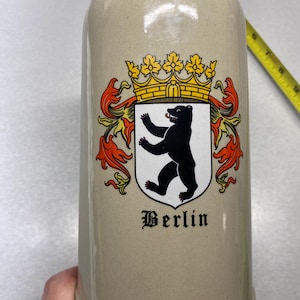May include: A beige ceramic beer stein featuring the Berlin coat of arms, which depicts a black bear standing on its hind legs beneath a yellow crown. The word Berlin is printed in black gothic-style lettering below the crest.