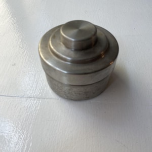 Könnte beinhalten: Ein zylindrischer Metallbehälter mit einem gestuften Deckeldesign. Das Objekt verfügt über eine glatte, gebürstete silberfarbene Oberfläche und einen kreisförmigen Knopf in der Mitte des Deckels. Es besteht aus einem Boden und einem Deckel.