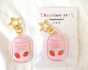 May include: Two pink and white strawberry cream keychain charms with gold star-shaped keychains. The charms are shaped like a can and have a cartoon strawberry on them. The charms are on a white background with a card that says "Kopitiam Art" and "@kopitiamart".