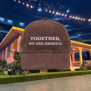 Puede incluir: Gorra de béisbol de pana marrón con el texto blanco «TOGETHER, WE ARE AMERICA». La gorra tiene una visera curva y una corona redondeada. El fondo presenta un edificio rosa y un estadio.