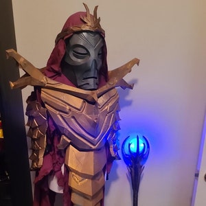 May include: A gold and black costume with a red cape and a blue glowing staff. The costume features a detailed dragon-like design and a mask with a crown.