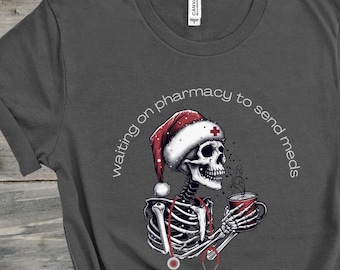 May include: Dark gray t-shirt featuring a skeleton wearing a Santa hat and holding a mug. The skeleton has a stethoscope around its neck and the text "waiting on pharmacy to send meds" is arched above the skeleton.