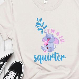 May include: Cream-colored t-shirt featuring a cartoon elephant squirting water from its trunk. The text "I'm a lil squirter" is printed in pink and blue. The elephant is holding an ice cream cone. The shirt is paired with blue jeans and black sneakers.