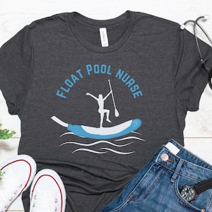 May include: Dark gray t-shirt with the text "FLOAT POOL NURSE" above a graphic of a person paddleboarding. The shirt is paired with denim shorts, white sneakers, and a stethoscope.