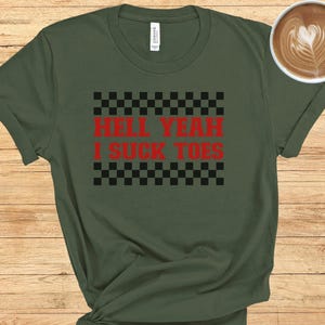 Puede incluir: Una camiseta verde con un patr&oacute;n de cuadros blanco y negro y el texto "Hell Yeah I Suck Toes" en rojo.
