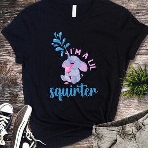 May include: Black t-shirt featuring a cartoon elephant squirting water and holding ice cream. The text "I'm a lil squirter" is in blue and pink. The shirt is paired with black sneakers and gray jeans.