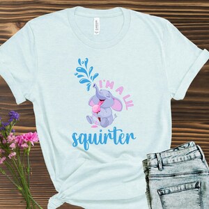 May include: Light blue t-shirt with a graphic of a cartoon elephant squirting water from its trunk. The text "I'm a lil squirter" is printed below the elephant in pink and blue. The shirt is paired with blue jeans.
