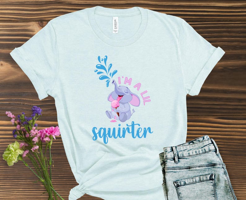 May include: Light blue t-shirt with a graphic of a cartoon elephant squirting water from its trunk. The text "I'm a lil squirter" is printed below the elephant in pink and blue. The shirt is paired with blue jeans.