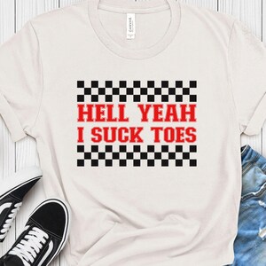 Puede incluir: Camiseta blanca con un patr&oacute;n de cuadros blanco y negro y el texto "Hell Yeah I Suck Toes" en rojo.