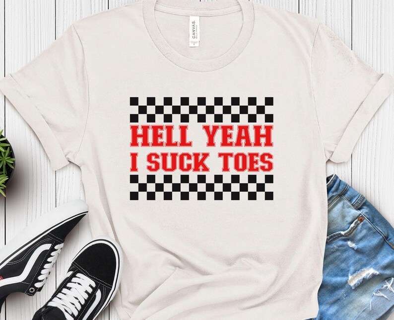 Puede incluir: Camiseta blanca con un patr&oacute;n de cuadros blanco y negro y el texto "Hell Yeah I Suck Toes" en rojo.
