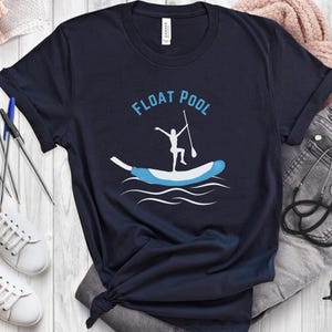 May include: Navy blue t-shirt with the words "FLOAT POOL" in light blue. The shirt features a graphic of a person paddleboarding on a blue and white float. White sneakers, pens, and other items are in the background.