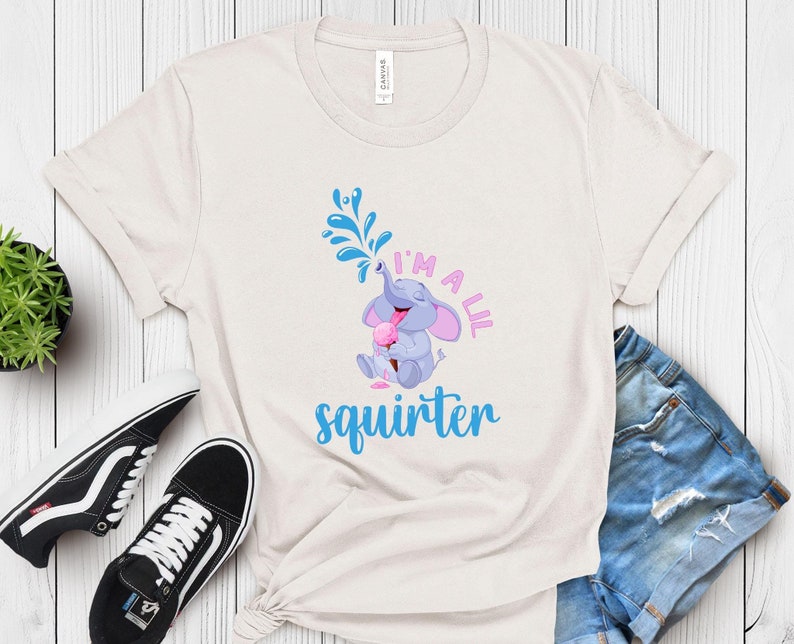 May include: Cream-colored t-shirt featuring a cartoon elephant squirting water from its trunk. The text "I'm a lil squirter" is printed in pink and blue. The elephant is holding an ice cream cone. The shirt is paired with blue jeans and black sneakers.