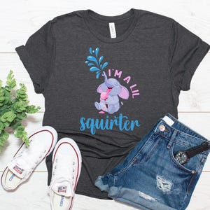 May include: Dark gray t-shirt with a playful graphic of a cartoon elephant squirting water, with the text "I'm a lil squirter" in blue and pink. The shirt is paired with denim shorts and white sneakers.