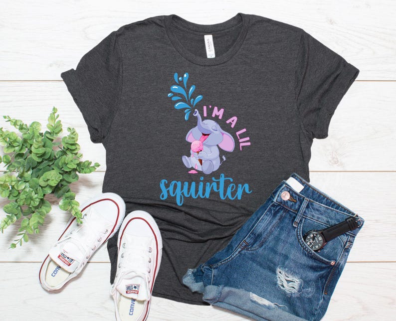 May include: Dark gray t-shirt with a playful graphic of a cartoon elephant squirting water, with the text "I'm a lil squirter" in blue and pink. The shirt is paired with denim shorts and white sneakers.