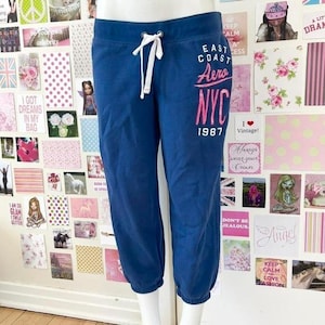Może przedstawiać: Granatowe spodnie dresowe z białym sznurkiem i różowym napisem "East Coast Aero NYC 1987". Te wygodne spodnie są odpowiednie do noszenia na co dzień.