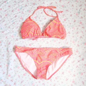 May include: A two-piece bikini set featuring a triangle halter top with thin straps and matching bikini bottoms. The swimwear is colored in shades of coral pink and yellow, decorated with a repeating geometric pattern across the fabric.
