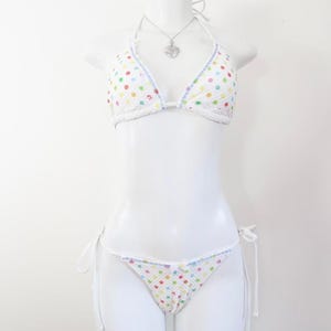 May include: White bikini set with colourful polka dots. The triangle top and string bikini bottoms feature a playful pattern of red, yellow, green, and blue dots. The edges are trimmed with white and light blue detailing.