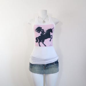 Pode incluir: Top branco com um painel rosa com uma silhueta preta de uma pessoa montando um unicórnio. O traje inclui uma minissaia jeans. Um colar de prata também é visível.