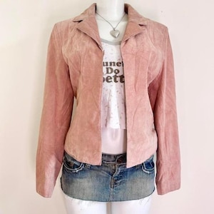 Op de afbeelding: Een lichtroze suède blazer met een kraag en lange mouwen. De blazer is open en onthult een wit t-shirt met de tekst "une Do ett" en een spijkerbroek mini-rok. Een zilveren hartvormige hangerketting is zichtbaar.