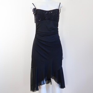 Puede incluir: Un vestido de noche negro con tirantes finos y un corpiño con lentejuelas. El vestido presenta una cintura fruncida y un dobladillo asimétrico con un efecto de capas. Una flor de tela adorna la correa izquierda. El vestido está en un maniquí.