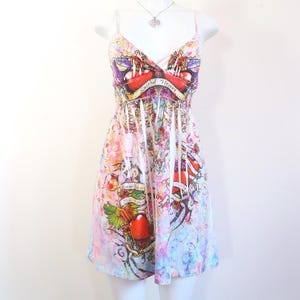 Puede incluir: Vestido sin mangas, hasta la rodilla, con estampado colorido estilo tatuaje. El vestido presenta un escote en V y tirantes finos. El estampado incluye corazones, rosas y las palabras "Sacred Heart" en una pancarta. El vestido se exhibe en un maniquí blanco.