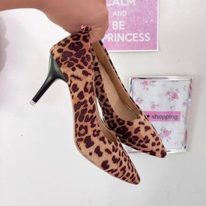 May include: A pair of pointed-toe high heels with a leopard print pattern. The heels have black stiletto heels with a small jewel detail at the base. The shoes are held up by a hand.