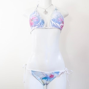 Puede incluir: Conjunto de bikini de dos piezas con estampado floral en tonos azules, rosas y blancos. El top halter y la braguita se exhiben en un maniquí blanco. También se ve un collar con colgante de corazón plateado.