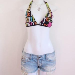 May include: A halter bikini top with the word "ROXY" in colorful letters, paired with light-wash denim shorts. The shorts have distressed details and a frayed hem. A silver heart pendant necklace completes the outfit. The ensemble is displayed on a white mannequin.