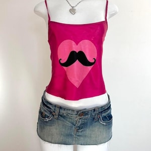 May include: A hot pink camisole with a light pink heart and a black mustache graphic. The camisole has thin straps and is paired with a denim mini skirt. A silver heart pendant necklace completes the outfit.