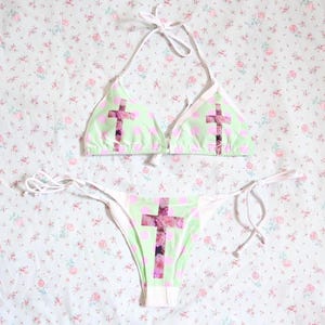 May include: A two-piece bikini set with a halterneck top and tie-side bottoms. The bikini is pale green with pink polka dots and a large cross design on the front. The cross design appears to be a floral pattern.