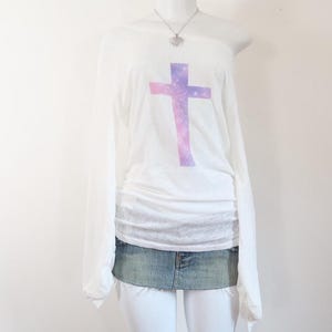May include: White long-sleeve top with a cross design in shades of purple and pink. The top is paired with a denim mini skirt and a silver necklace with a heart pendant. The top has an off-the-shoulder design.