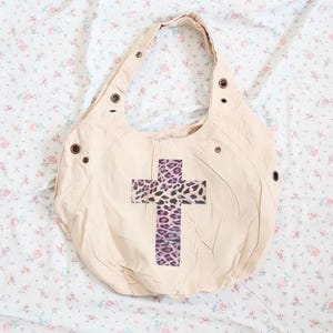 May include: Beige canvas hobo bag with a cross design. The cross features a leopard print pattern with purple accents. The bag has a single shoulder strap and metal grommets along the top edge.