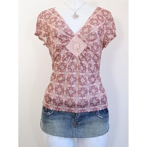 May include: A short-sleeved, V-neck top with a floral pattern in shades of pink and brown. The top features a decorative square embellishment at the neckline and is paired with a denim mini-skirt. A silver necklace with a heart pendant is also visible.