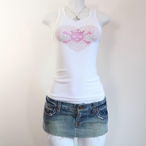 Może przedstawiać: Biały prążkowany top na ramiączkach z różową grafiką w kształcie serca z czaszką i skrzyżowanymi piszczelami. W połączeniu z dżinsową mini spódniczką. Srebrny naszyjnik z wisiorkiem w kształcie serca dopełnia stylizację.