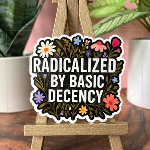 May include: A sticker with the text "RADICALIZED BY BASIC DECENCY" in a white, bold font. The text is surrounded by a black floral design with colorful flowers in shades of pink, orange, blue, and white. The sticker is displayed on a small gold easel.