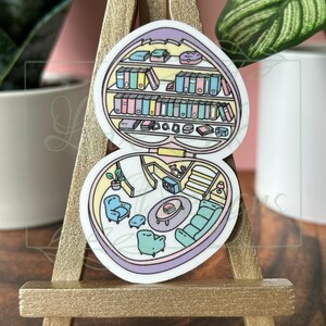 May include: A sticker of a heart-shaped house with a bookshelf, a couch, a chair, and a coffee table. The house is open and the interior is visible. The sticker is on a wooden easel.